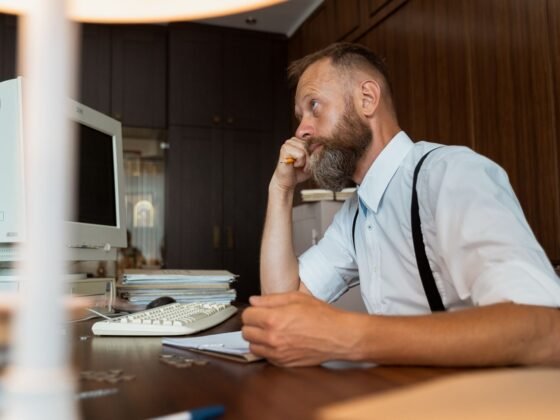 Un homme mûr à la barbe rousse et grise, en réflexion à son bureau, une main contre le visage.